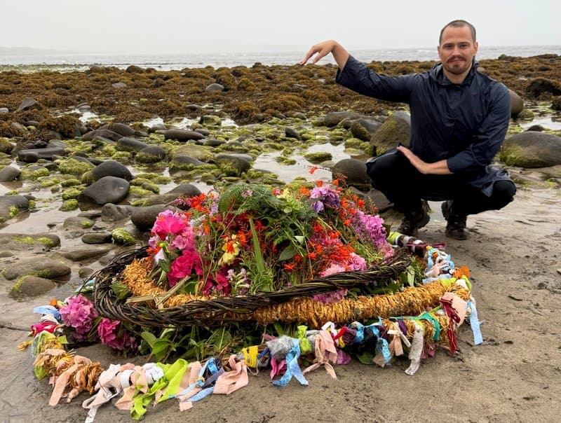 Russell Patrick Brown in a squatting dance pose on a beach with large rocks next to ceremonial Lúnasa wreaths